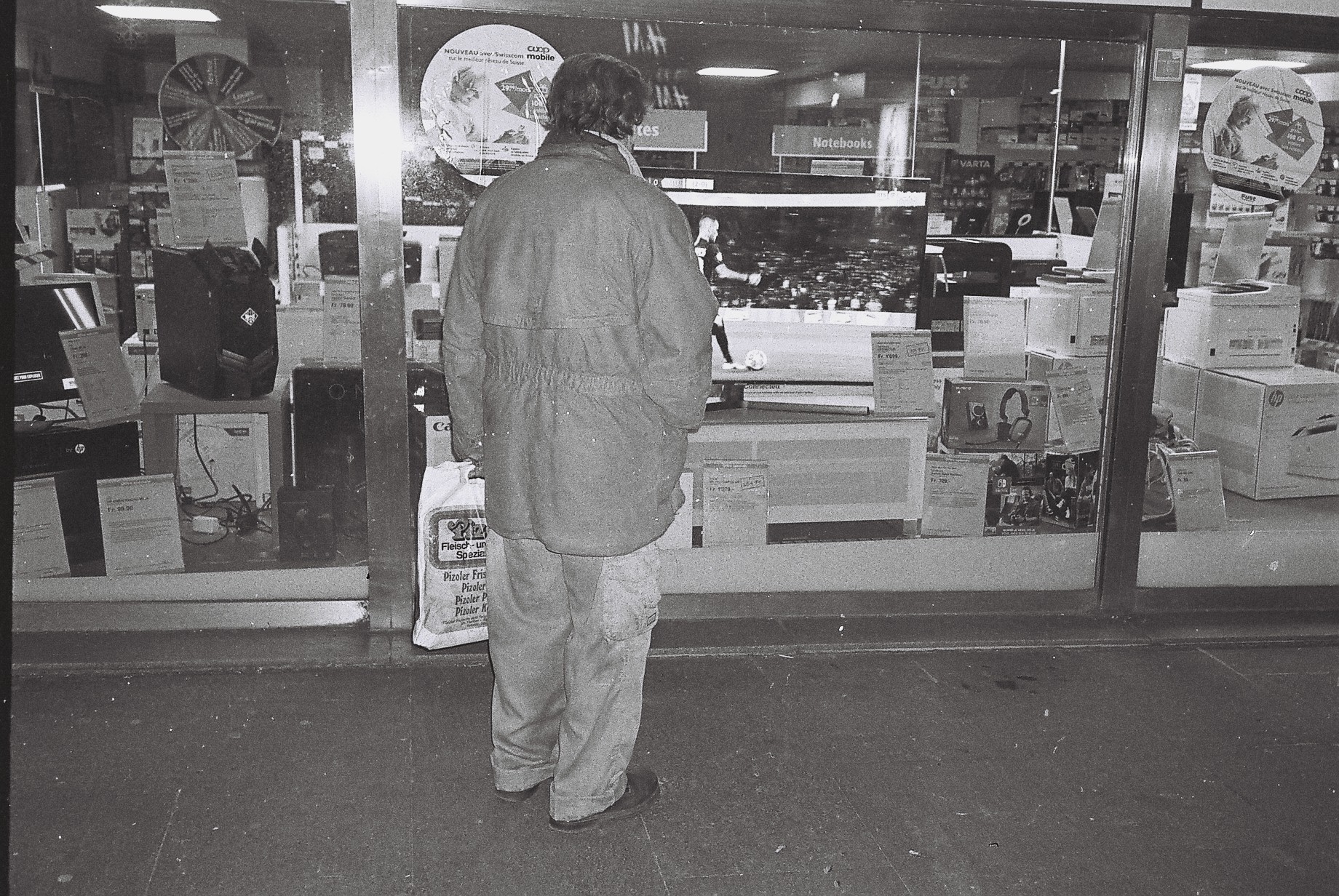 A black and white photo of a man carrying a shopping bag and looking through a shop window at a tv showing football