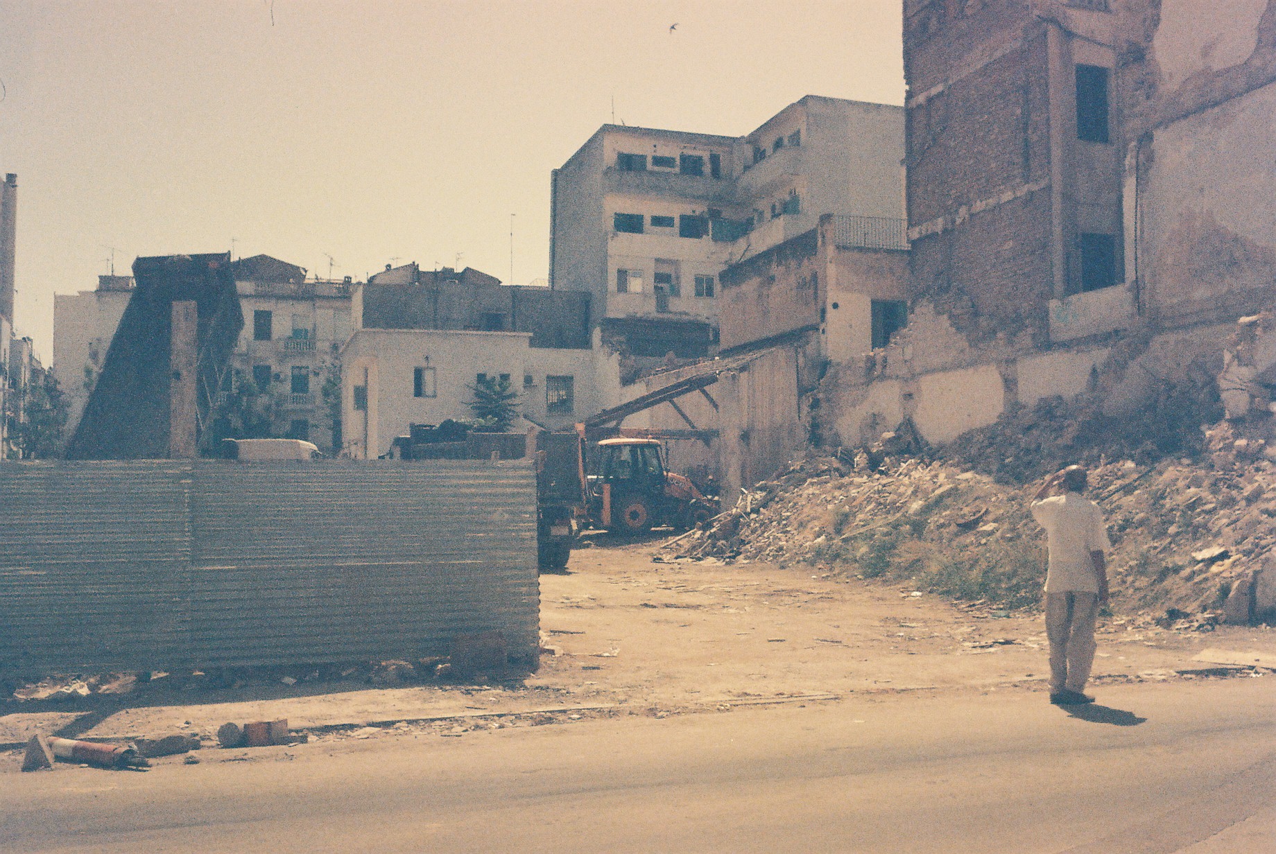 A man is loose trousers and a short sleeved shirt standing in front of a building site, looking at it, shading his eyes